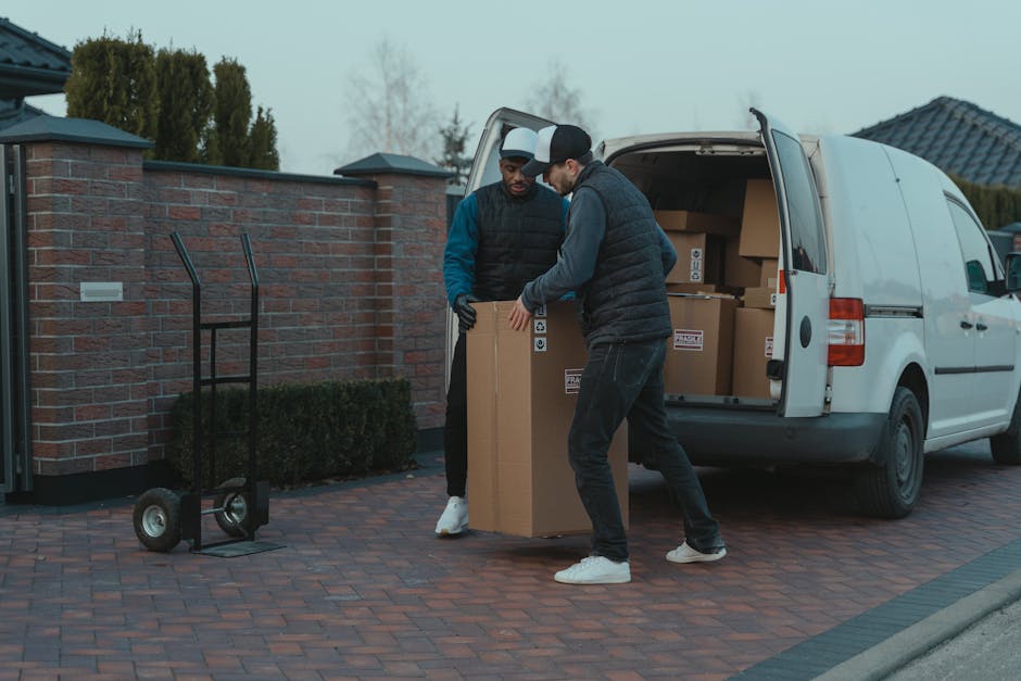 Two men are engaged in a house relocation task outdoors, loading a cardboard box into the back of a white moving van parked on a paved driveway. One man, wearing a black vest and cap, is holding the box steady, while the other, dressed in a dark quilted jacket and black cap, is assisting by guiding the box into the vehicle. A black hand truck with two wheels and a handle rests nearby on the brick pavement, ready for transport. The open van reveals several packed boxes stacked inside, some sealed with packing tape. The background features a brick wall with a gate, hedges, and residential houses with tiled roofs, under a cloudy sky. The scene captures the logistics of furniture transport and packing during a home relocation, with natural lighting highlighting the action of loading items for moving services provided by [COMPANY_NAME].