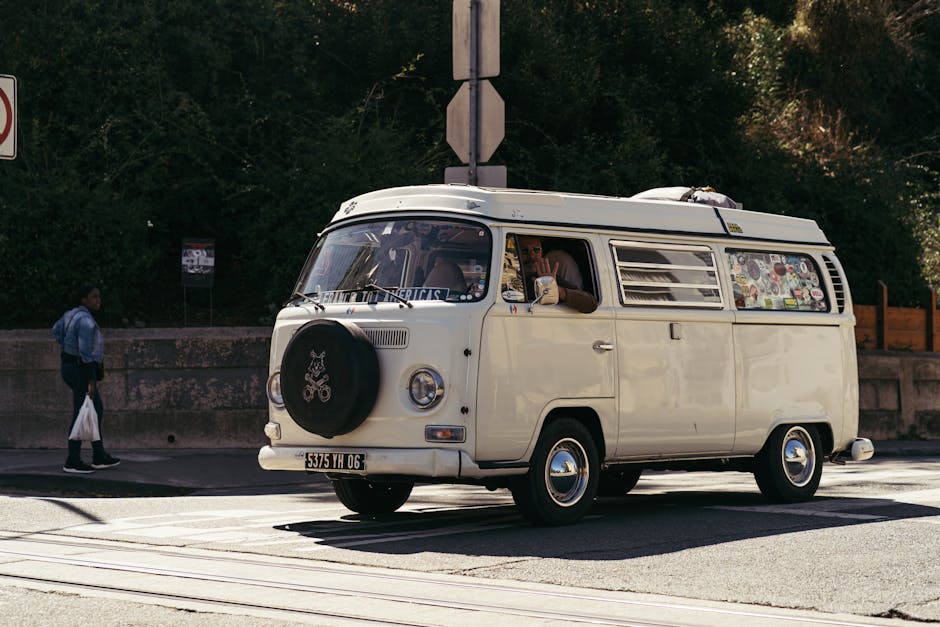 A white moving van marked with the text 'MOVING COMPANY LOCAL & LONG DISTANCE' is parked on the street in front of an urban brick building with large ground-floor windows and two arched windows on the upper floor. The driver, wearing sunglasses and a dark jacket, is sitting in the open driver's side window, holding the steering wheel and looking towards the camera. The van is positioned close to the curb, with its rear section aligned for easy loading or unloading of furniture, boxes, and packing materials. In the background, there are other trucks and vehicles, including a yellow truck, along with a tree and clear blue sky, indicating daytime. This scene reflects a home relocation or furniture transport process involving loading or preparing for a move, as managed by [COMPANY_NAME], a professional removals service specializing in house moves and logistics.