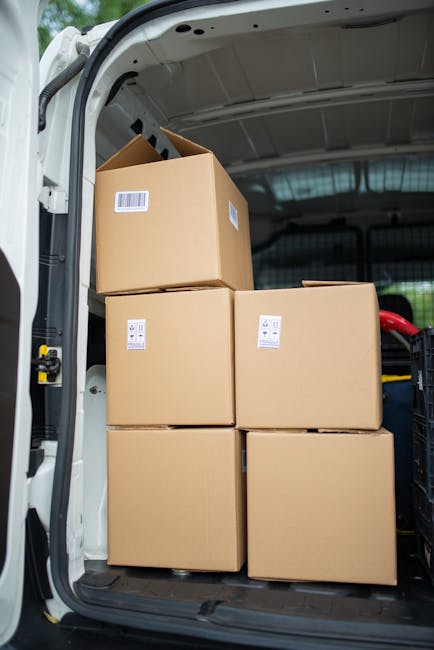 Inside the rear of a moving van during a home relocation, with several cardboard boxes of varying sizes stacked near the open tailgate, some sealed with packing tape and others uncovered, ready for tr