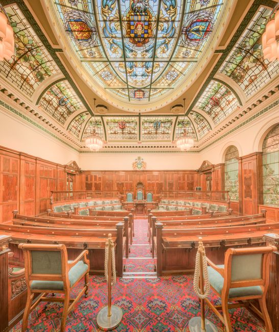The image depicts a large, ornate parliamentary chamber illuminated by warm lighting, featuring rows of wooden desks and chairs arranged in a semi-circular pattern facing the central speaker's podium.