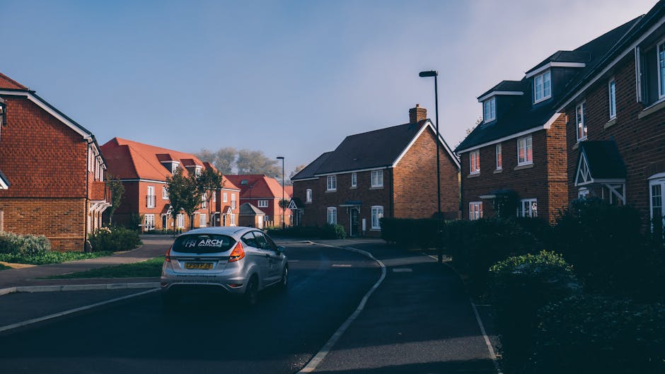 A residential street in Arkley EN5 featuring several red-brick and dark-brick semi-detached houses with pitched roofs and white-framed windows, set along a curving road with a designated parking lane on the right. A silver hatchback van labeled 'Man with Van Arkley' is parked partially on the pavement and the street, positioned near the curbside. The van is in the process of loading or unloading household items, indicated by nearby cardboard boxes, a small wooden table, and protective moving blankets spread on the ground and inside the vehicle. The environment is lit with natural daylight, casting mild shadows, and a streetlamp stands on the sidewalk, guiding the way for moving logistics. The scene captures the typical home relocation activity with the presence of packing materials, furniture, and transport equipment being prepared for a house move, aligning with services offered by Man with Van Arkley focused on removals and furniture transport in Arkley.