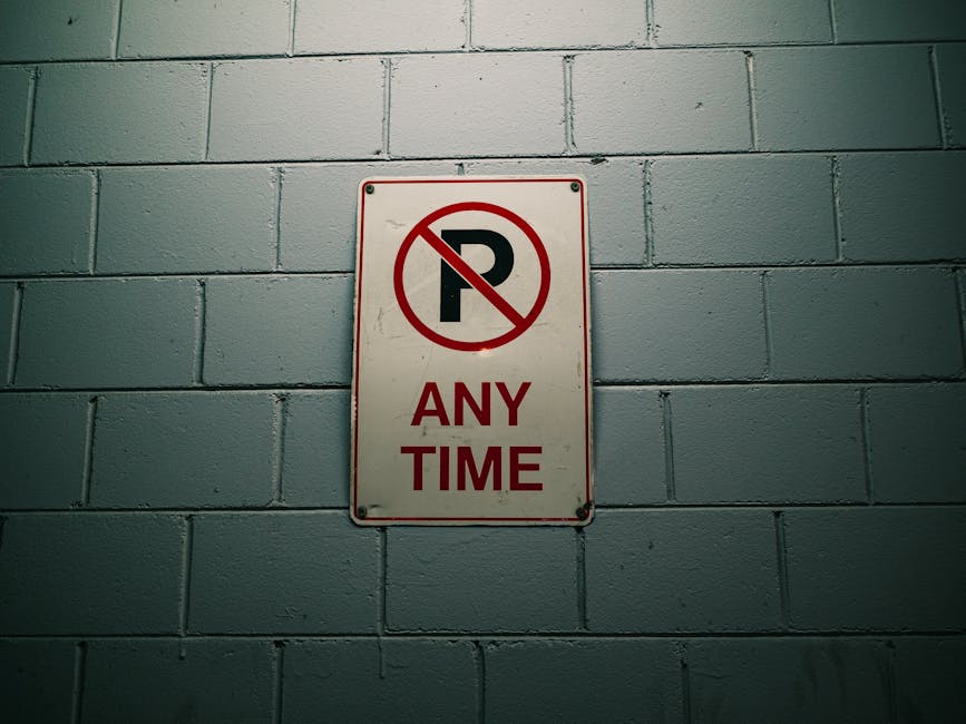 A rectangular metal 'No Parking Any Time' sign with a red circle and slash over a black letter 'P' is mounted on a grey painted brick wall indoors. The wall has visible horizontal mortar lines, and the sign is secured with four screws, one at each corner. The area is evenly lit, showing the sign clearly. This setting suggests a confined indoor environment, possibly a parking garage or storage area, where parking restrictions are in force. The context of house removals and furniture transport may relate to managing parking rules during a home relocation or loading process, with Man with Van Arkley providing removal services. The sign's message indicates that parking is prohibited at all hours, which could be relevant during logistics planning for moving or unloading at the property located in Arkley EN5, supporting best practices to avoid parking fines or obstructions during a move.