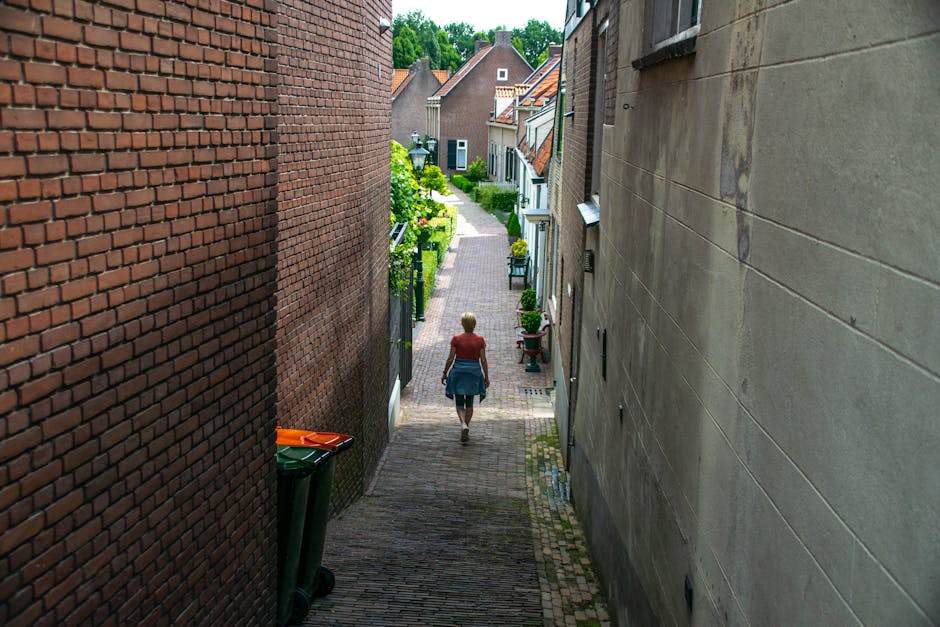 A woman walking down a narrow residential alleyway between two rows of houses, with brick walls on the left and a stone façade on the right. The alley is paved with bricks and cobblestones, leading to a landscaped street with houses and greenery in the background beneath a partly cloudy sky. The woman wears a red top, jeans, and a skirt, and appears to be heading towards a house with potted plants and decorative elements outside. This scene captures the process of home relocation, potentially involving furniture transport and moving logistics managed by Man with Van Arkley, with visible elements like the alleyway environment and the general context of moving within Arkley's neighbourhood.