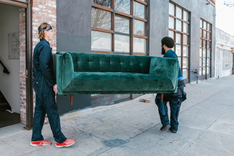 Two men are engaged in a furniture transport activity outside a building on a city street. One man with long hair, wearing a navy blue jacket, navy pants, and red sneakers, stands on the sidewalk, observing the process. The second man, with a beard, dressed in dark clothing, is carrying a large, green velvet sofa with wooden legs, holding it securely with both hands. The sofa is positioned horizontally, with its backrest facing the building's exterior wall, which features large windows framed by reddish-brown wooden supports. The building has a dark exterior surface, and the sidewalk is made of concrete. The scene indicates an outdoor home relocation or furniture moving process, likely coordinated by a professional removals service such as Man with Van Arkley, as part of a house move or furniture transfer operation. The natural lighting suggests daytime conditions, and the surroundings include adjacent buildings and a street with closed garage doors in the background.