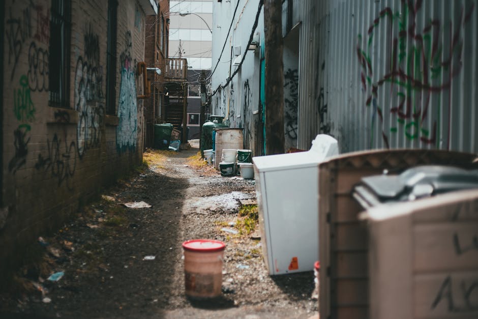 A woman walking down a narrow residential alleyway between two rows of houses, with brick walls on the left and a stone façade on the right. The alley is paved with bricks and cobblestones, leading to a landscaped street with houses and greenery in the background beneath a partly cloudy sky. The woman wears a red top, jeans, and a skirt, and appears to be heading towards a house with potted plants and decorative elements outside. This scene captures the process of home relocation, potentially involving furniture transport and moving logistics managed by Man with Van Arkley, with visible elements like the alleyway environment and the general context of moving within Arkley's neighbourhood.