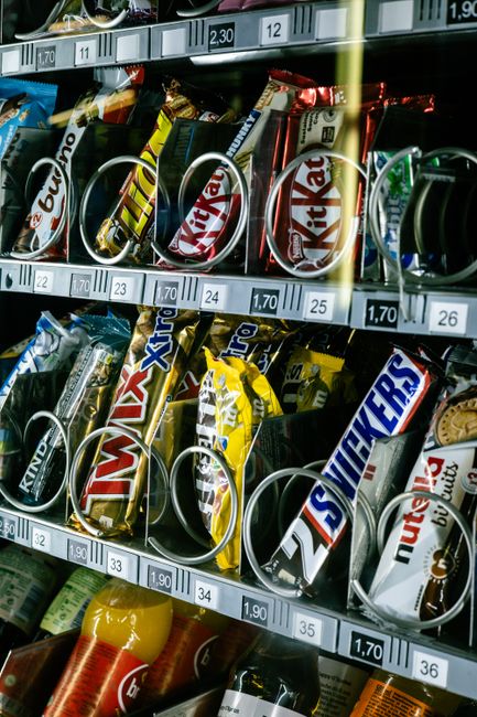 Close-up image showing a vending machine filled with various snack bars, including Kit Kat, Snickers, Twix, and Nutters, organized in metal wire compartments with visible price labels. The vending mac