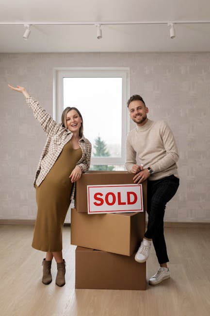 Two individuals, a woman and a man, stand inside an empty room with a large window behind them, showing a cloudy sky and trees outside. The woman, dressed in a beige checkered shirt over a mustard dress, has her left arm raised in celebration and is smiling at the camera. The man, wearing a light grey sweater and dark trousers, is holding a cardboard box labeled 'SOLD' on top of two other stacked boxes, one of which is partially opened, indicating recent house sale and moving activities. The room has light-colored walls and a wooden floor, with a ceiling fitted with white track lighting. The scene captures a moment of home relocation success, likely involving furniture transport, packing, and moving logistics, which is relevant to professional removals services by Man with Van Arkley. The overall setting suggests a successful property sale completed, ready for the next stage of the moving process.