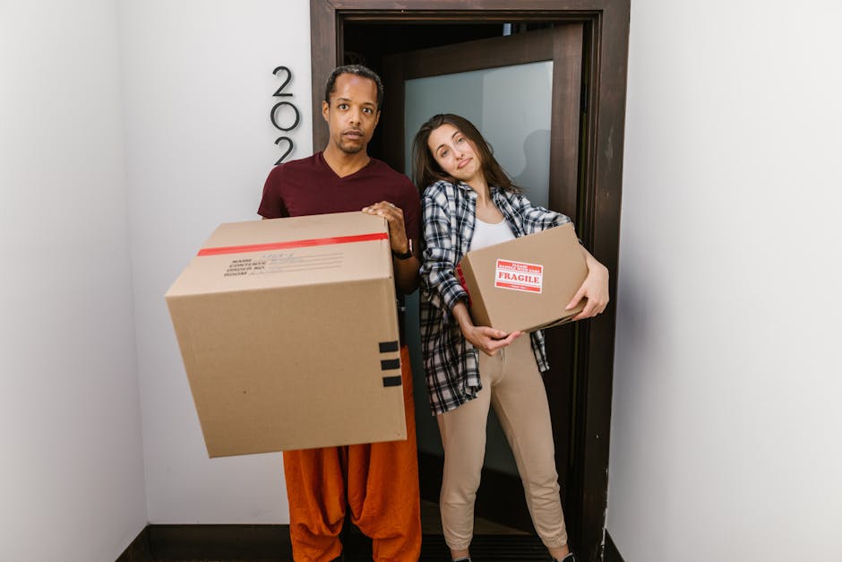 A man with a cardboard box standing beside a woman holding another packed cardboard box at the doorway of a property during a house removal. The man is wearing orange trousers and a maroon t-shirt, while the woman sports beige trousers and a blue plaid shirt. Both are inside a narrow corridor, with the doorway leading to an interior room behind them. The wooden frame door is open, revealing a frosted glass panel. The boxes are labeled with fragile and mailing information, indicating careful packing for a home relocation. This scene reflects a loading process, with the man with van Arkley team preparing to transport household belongings, including furniture and packed boxes, as part of a furniture transport or moving service. The setting is well-lit, with the focus on the handling and organization of items ready for transport to a new residence, illustrating the logistical aspect of house removals.